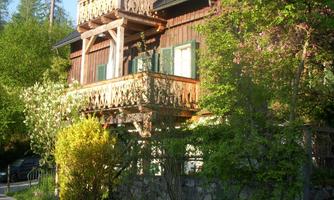 A charming wooden house with a balcony, surrounded by lush greenery. Tall trees stand in the background and the sky is clear.