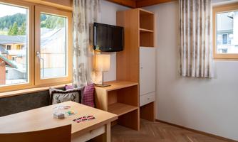 A cozy room with two windows and a desk. A small chest of drawers and a television are also available.