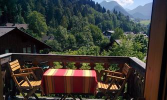 A beautiful balcony with wooden chairs and a table covered with a colorful tablecloth. In the background, green trees and mountains can be seen.