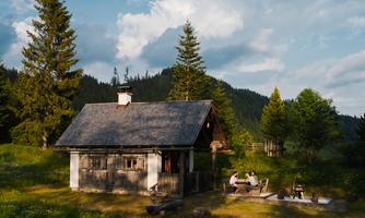A small wooden cabin surrounded by green meadows and tall trees. In the foreground, people are sitting by the campfire and enjoying nature.