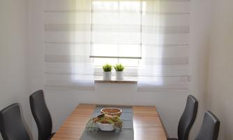 A bright dining area with a wooden table and four chairs. A decorative cloth is laid on the table and small plants are on the windowsill.