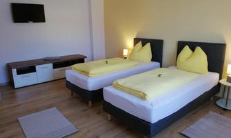 A modern bedroom with two single beds and yellow cushions. In the room, there is a television and a desk.