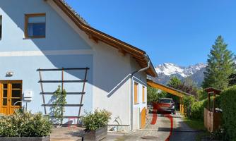 A modern house with a green outdoor area and a view of the mountains. A car is parked next to the house under a canopy.