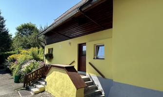 A yellow house with a wooden roof and a staircase. It is surrounded by colorful flowers and greenery.