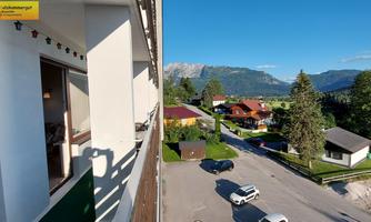 A beautiful view from a balcony overlooking a quiet residential area with houses and trees. In the background, mountains are visible under a clear blue sky.