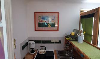A cozy kitchen with a stove, sink, and a window. A colorful picture hangs on the wall, and the countertop is decorated with kitchen utensils.