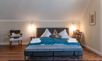 A cozy bedroom with a large bed and colorful cushions. There are chairs and soft lighting on the walls, creating a relaxed atmosphere.