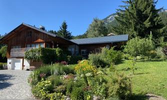 A cozy wooden house surrounded by a blooming garden. In the background, trees and mountains can be seen.