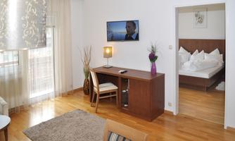 A cozy room with wooden flooring and a desk. In the background, a bedroom with a bed and white pillows is visible.