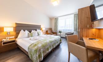 A modern hotel room with a double bed and bright wooden accents. There is a desk, two chairs, and large windows that provide plenty of daylight.