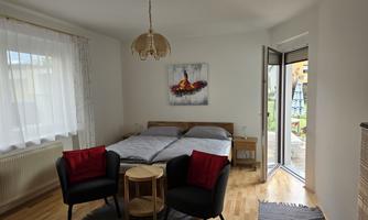 A cozy bedroom with a double bed and two black armchairs. Large windows let in plenty of light, and a door opens to the garden.