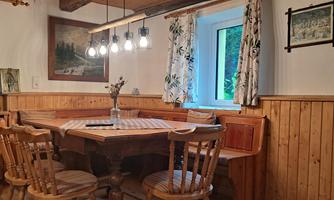 A cozy dining area with a round wooden table and wooden chairs. Large windows let in plenty of daylight and the walls are clad with wooden panels.