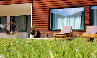 A modern house with wooden cladding and large windows. In the foreground, two loungers are visible on a green lawn.