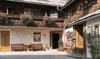 A picturesque courtyard with two traditional houses and colorful flowers. The entrance doors are inviting and the sky is clear.