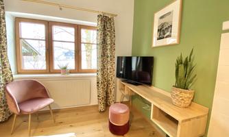 A cozy room with green wall color, a television, and wooden furniture. Large windows let in a lot of light, and the room is decorated with plants.