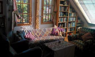 A cozy living room with large windows and sunshine streaming through the blinds. Colorful cushions are on the sofa, and in the background, there is a bookshelf.