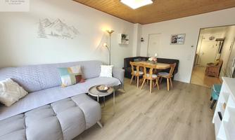 A cozy living room with a gray couch and a wooden table. There are bright walls and a wooden ceiling, creating an inviting atmosphere.