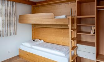 A simple room with a wooden bunk bed and a shelf. The walls are neutral and there are curtains at a window.