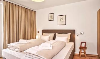 A cozy bedroom with two single beds and a warm, neutral color palette.  
Large windows with curtains provide plenty of daylight and a pleasant atmosphere.