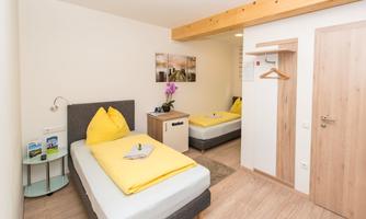 A cozy room with two beds and yellow pillows. There is a nightstand, a wardrobe, and a window with plenty of natural light.