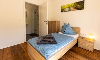 A cozy bedroom with a modern bed and a blue duvet. In the background, a bathroom is visible and a decorative picture on the wall.