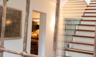 A modern hallway with a wooden staircase. Through a grid window, the view of the area behind the staircase can be seen.