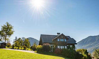 A beautiful, modern house on a green meadow. In the background, there are mountains and a blue sky with sunlight visible.