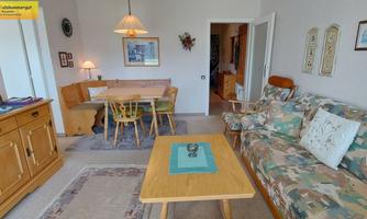 A cozy living and dining room with a sofa, a table, and chairs. The walls are tastefully decorated, and there is plenty of natural light.