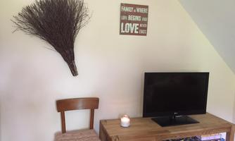 A cozy living room with a television and a wooden shelf. A decorative branch fork hangs on the wall along with a sign featuring a family motto.