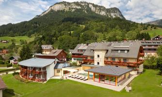 A picturesque hotel building in a rural setting with a large garden and a view of the mountains. The landscape is green and inviting, perfect for relaxation and outdoor activities.