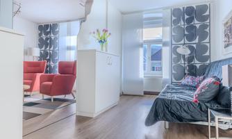 A modern furnished living space with red armchairs and a sofa bed. The large windows provide plenty of daylight.