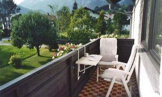 A cozy balcony with a white chair and table. In the background, green trees and mountains can be seen.