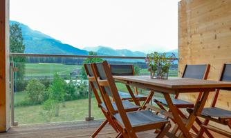 A cozy terrace with a wooden table and chairs. In the background, gentle mountains and a green landscape can be seen.