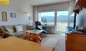 A cozy living room with a comfortable seating area and a wooden table. Large windows offer a view of the mountains and plenty of natural light.