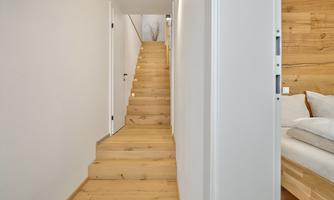 A bright hallway with wooden stairs leads to a bedroom. The walls are in neutral colors and the floor is made of wood.