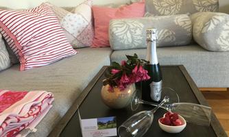 A cozy living room with a gray couch, colorful cushions, and a table. On the table are champagne, glasses, a flower vase, and fresh raspberries.