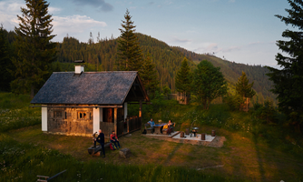 A cozy cabin in nature, surrounded by trees and meadows. People are sitting relaxed outside, enjoying the evening atmosphere.