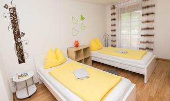 A bright bedroom with two single beds and yellow pillows. The room is simply furnished and has large windows with curtains.