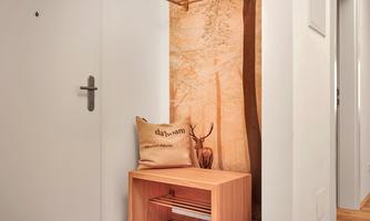 A modern entrance area with a wooden stool and a cushion. In the background, there is a wall decoration featuring a landscape motif.