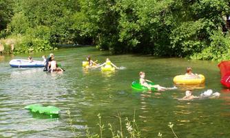 People are swimming and splashing in the clear water of a river. Some are using inflatable tubes in various colors.