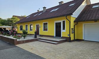 A modern, yellow house with a landscaped front garden. The driveway is paved with cobblestones and provides space for vehicles.