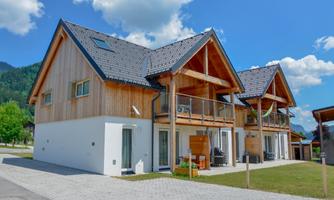 A modern two-story house with wooden cladding and balconies. The surroundings are green and rural, with a blue sky and clouds.