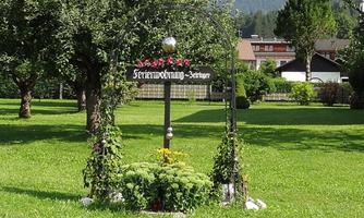 A beautiful entrance with a decorative arch and colorful flowers stands in a green meadow. In the background, houses and mountains are visible.