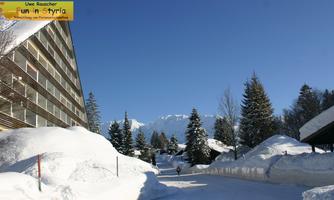 A snow-covered path with high snowdrifts and green fir trees. In the background, snow-covered mountains and a blue sky are visible.
