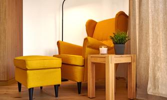 A cozy living area with a yellow armchair and a matching footstool. A small wooden table with a houseplant and a candle adds to the relaxed atmosphere.
