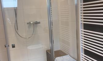 A modern shower with glass partition and gray tiles. In the background, radiators and a towel can be seen.