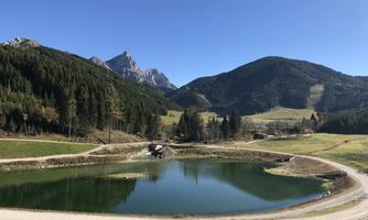 A tranquil landscape with a clear pond, surrounded by mountains and forests. The sky is blue and the sun is shining.