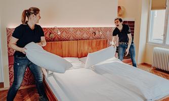 Two women are making a bed in a cozy room. They are holding pillows and blankets as they lay down the bed linens.