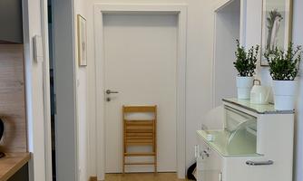 A bright hallway with wooden flooring and a white wall. Plants in pots are standing against the wall, and a small chair is visible.