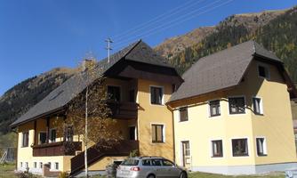A two-story yellow house in the midst of a mountain landscape. Next to the house is a car, and the sky is clear and blue.